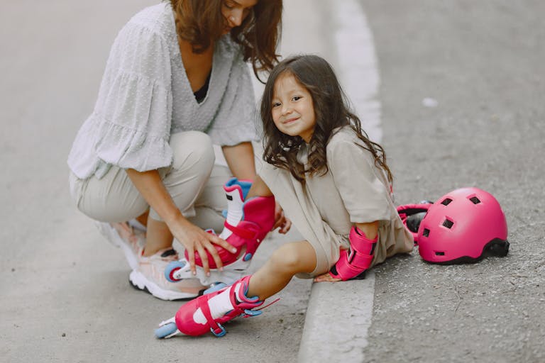 A young girl wearing pink rollerblades sits on a curb while her mother assists her. Outdoors setting.